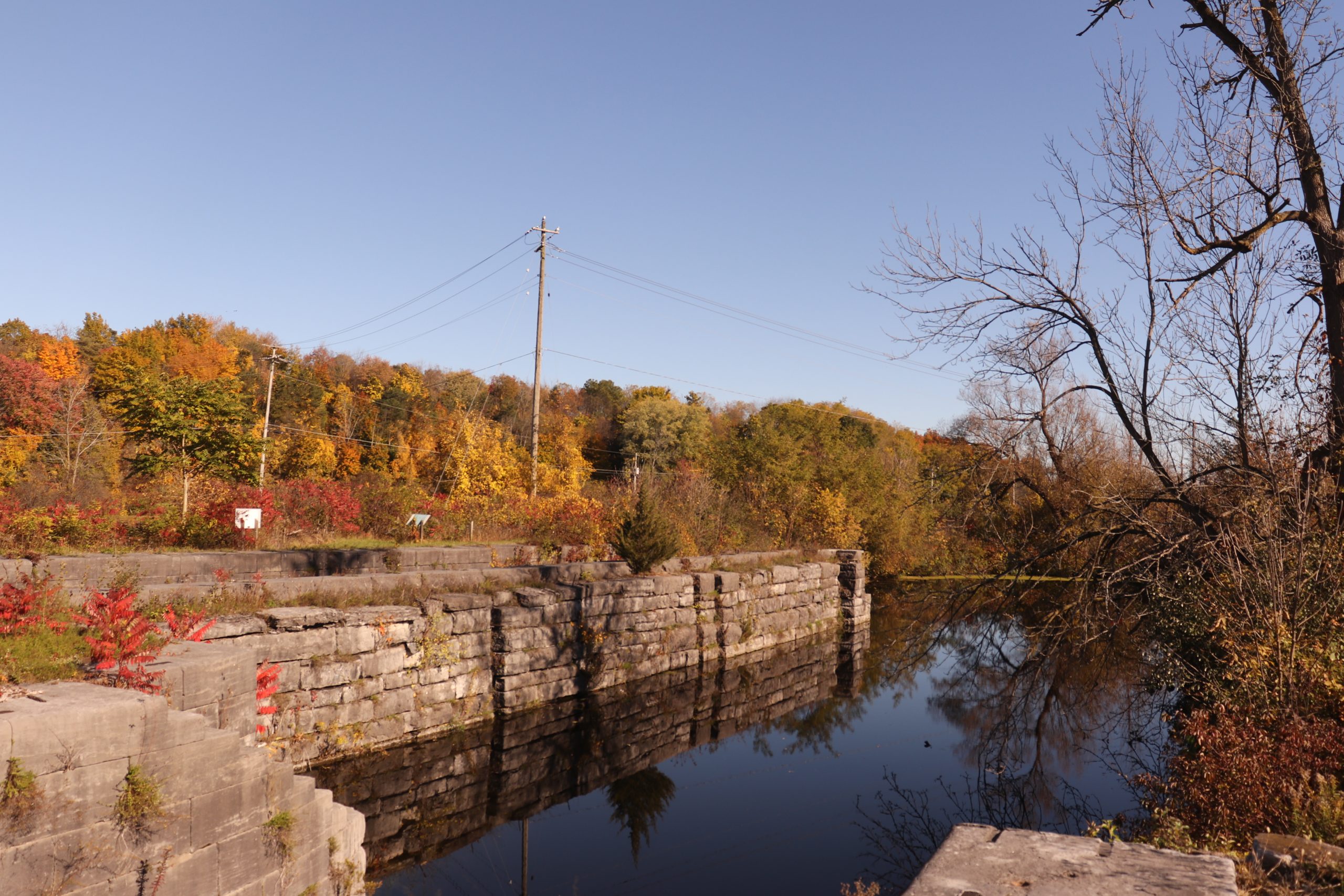 Fall foliage decorates the original Erie Canal and the Erie Barge Canal behind the Poorhouse Grocery Canal Store, now the private residence of Allyn Hess Perry, off of Dry Dock Road in Lyons, New York. Markers on the Enlarged Erie Canal Lock 56 Trail can be seen through the overgrown shrubbery | Photo by Julia Carden
