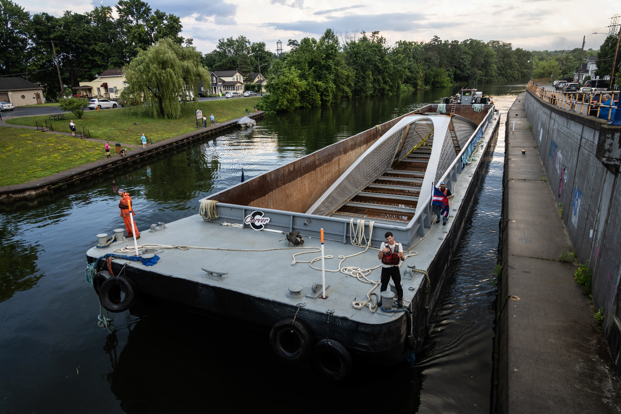 The CMT Kelly barge, pushed by CMT Otter tugboat and carrying a section of the Ralph Wilson Park Bridge, approaches Lock 24 in Baldwinsville, New York on Wednesday, July 10, 2024. The barge, one of two, is traveling the Erie Canal toward its final destination in Buffalo, New York. The second barge, CMT Hoffman, remained docked at Lock 23 in Brewerton Wednesday night due to higher than anticipated water levels, according to Carver Marine Companies. Photo by Kayla Breen