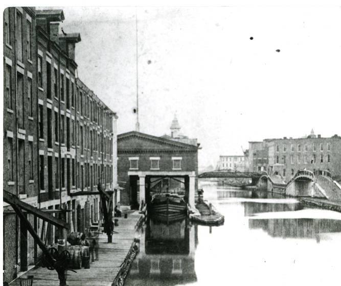 Looking West on the Erie Canal, now Erie Boulevard. To the left of the Weighlock Building are warehouses with loading rigging, to the right is the Oswego Canal. A canal boat has been drawn into the lock, waiting for the lock gates to be closed and water drained out, so a tollmaster can weigh it on a cradle that is attached to a scale.